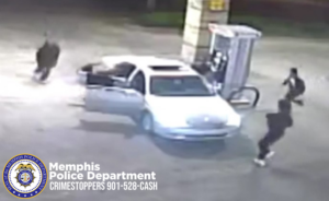 A grainy surveillance-style image shows a light-colored sedan parked at a gas pump at night. The front passenger door of the vehicle is open. Three individuals wearing dark clothing are moving rapidly around the vehicle: one near the open passenger door, one near the front of the car, and one farther out in the background on the left side of the image. A gas pump and its overhead structure are visible to the right of the car. In the lower-left corner, there is a Memphis Police Department badge logo with white text reading “Memphis Police Department CRIMESTOPPERS 901‑528‑CASH.”