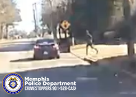 A vehicle is stopped on a residential street near a crosswalk sign. A person is moving across the grass toward the road on the right side of the image. Trees and houses line the background. In the lower-left corner, there is a Memphis Police Department Crimestoppers graphic with the phone number 901‑528‑CASH.