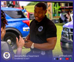 An outdoor community event scene shows an individual wearing a black polo shirt with an embroidered Memphis Police Department emblem on the chest and a duty belt with a badge. The individual is engaged in conversation with another person in the foreground who is gesturing with both hands. Behind them, brightly colored vehicles, including a blue police SUV and another blue vehicle, are parked on a grassy area. Additional people and vendor tents or booths are visible in the background. At the bottom of the image, a banner displays the Memphis Police Department seal, the text “Memphis Police Department – Established in 1827,” and the website joinmpd.com along with several social media icons.