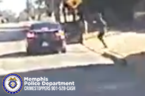 A vehicle is stopped on a street in a residential area. To the right of the vehicle, a person is moving across a grassy area near the curb. A crosswalk warning sign is posted on a pole behind the person. Trees and houses line the background. In the lower-left corner, there is a Memphis Police Department Crimestoppers graphic with the phone number 901‑528‑CASH.