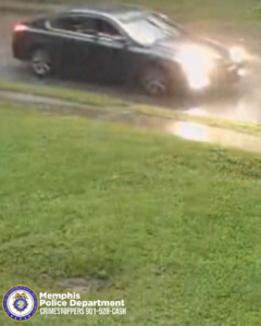 A dark-colored sedan drives on a wet, reflective road near a grassy area. Its headlights are on, suggesting rainy or damp conditions. In the lower-left corner, a Memphis Police Department Crimestoppers graphic displays the text “901‑528‑CASH.”