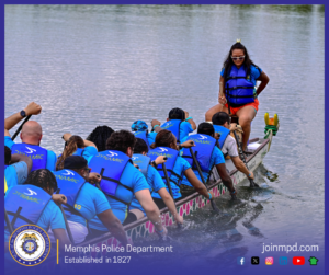 A long dragon boat filled with a paddling team is moving across a calm body of water. The team members, all wearing matching bright blue shirts and blue life vests labeled “DYNAMIC,” are seated in rows and paddling in unison. At the front of the boat sits an individual wearing a life vest and holding a steering paddle while facing the paddlers. The dragon boat has a decorative dragon head at the front, painted in green and yellow. The background consists of smooth water extending across the entire scene. Along the bottom edge of the image, a banner displays the Memphis Police Department seal, the text “Memphis Police Department – Established in 1827,” the website joinmpd.com, and several social media icons.