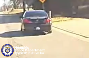An image shows the rear view of a dark-colored car driving on a residential street in daylight. The vehicle is positioned near the center of the road, and sunlight creates bright reflections on its surface and the pavement. A crosswalk warning sign is visible ahead on the right side of the road. The right side of the image shows a grassy area and trees casting shadows. In the lower-left corner, a Memphis Police Department Crimestoppers graphic appears with the phone number 901‑528‑CASH.