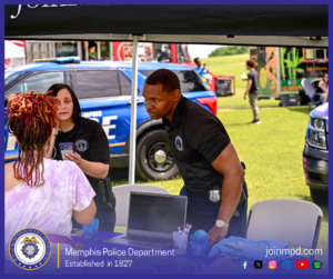 An outdoor community event scene shows two Memphis Police Department personnel standing under a canopy tent while interacting with a visitor who is holding a mobile phone. On the table beneath the tent, there is a laptop, informational materials, cups, and promotional items. Behind the group, a blue Memphis Police Department vehicle is parked on a grassy area. Additional people, food trucks, and vendor booths are visible in the background across an open field. At the bottom of the image, a banner displays the Memphis Police Department seal, the text “Memphis Police Department – Established in 1827,” the website joinmpd.com, and several social media icons.