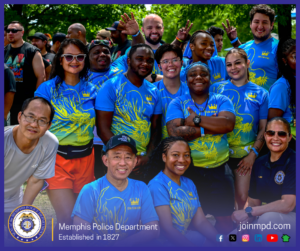 A large group of people is gathered outdoors in a park-like setting, posing closely together for a group photo. Most individuals are wearing matching blue shirts with bright yellow dragon designs and text that appears to say “Dim Sum King.” Several people are making hand gestures such as raised arms or peace signs. The group is standing and crouching in multiple rows, with trees and other event participants visible in the background. At the bottom of the image, a banner features the Memphis Police Department seal, the text “Memphis Police Department – Established in 1827,” the website joinmpd.com, and various social media icons.