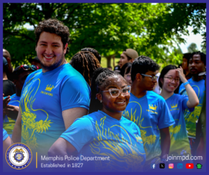 A group of people are standing outdoors in a park-like setting with trees providing shade in the background. Several individuals in the foreground are wearing bright blue shirts featuring a large yellow dragon graphic and the text “DIM SUM KING.” Some people stand closely together, while others appear slightly turned or engaged with those around them. Additional people can be seen in the background, some wearing casual clothing and holding personal items. At the bottom of the image, a banner displays the Memphis Police Department seal, the text “Memphis Police Department – Established in 1827,” the website joinmpd.com, and various social media icons.