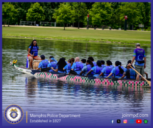 An individual is seated at the front of a dragon boat on calm water. The individual is wearing a blue and black life vest over a blue athletic shirt with a yellow graphic, along with bright orange shorts. One hand is holding a wooden drumstick or paddle handle, while the other hand is positioned near the side of the body. The water stretches across the background with grassy shoreline visible in the distance. Along the bottom of the image, a banner displays the Memphis Police Department seal, the text “Memphis Police Department – Established in 1827,” the website joinmpd.com, and several social media icons.