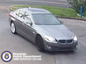 A surveillance-style image shows a gray sedan, appearing to be a BMW, driving on a paved area near a street. The vehicle’s headlights are on, and the front left wheel appears darker than the rear wheel. A grassy roadside area is visible in the background. In the bottom-left corner of the image, there is a Memphis Police Department badge graphic with text that reads: “Memphis Police Department CRIMESTOPPERS 901-528-CASH.”