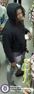A person wearing a dark hooded sweatshirt and light-colored pants is standing inside a store aisle. The individual is holding small boxed items in both hands. Nearby shelves are stocked with various brightly packaged snacks. Another person is partially visible to the left side of the frame. The floor appears to be smooth and reflective. In the bottom-left corner, there is a Memphis Police Department badge graphic with text reading: “Memphis Police Department CRIMESTOPPERS 901-528-CASH.”
