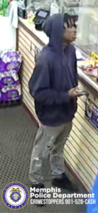 A person wearing a dark hooded sweatshirt is standing at a store counter. The individual is holding a phone or similar handheld device in one hand while facing the counter area, which has various small items displayed near the edge. The person is also wearing light-colored cargo-style pants and dark footwear. To the left, several stacks of purple-packaged products are arranged on shelves. The floor is covered with a textured, dark-colored mat. At the bottom-left corner of the image is the Memphis Police Department badge graphic with text that reads: “Memphis Police Department CRIMESTOPPERS 901-528-CASH.”