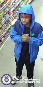 A person wearing a bright blue hooded jacket with darker blue sections stands inside a store aisle. The hood is pulled up, and the individual’s hands are clasped together at chest level. A black shirt is visible under the jacket, and the person is wearing dark pants with a vertical stripe on the side, along with athletic-style shoes. To the left, shelves are filled with assorted packaged snacks in various colors. The floor appears to be smooth and light-colored. At the bottom-left corner, a Memphis Police Department badge graphic is displayed with the text: “Memphis Police Department CRIMESTOPPERS 901-528-CASH.”