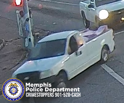 A white pickup truck with a damaged or missing tailgate is captured in motion at an intersection. The truck is turning right, and the front end is slightly blurred due to movement. A traffic signal pole and crosswalk lines are visible nearby. Another white vehicle with headlights on is partially visible behind the truck. In the lower left corner, there is a Memphis Police Department badge graphic and text that reads “Memphis Police Department CRIMESTOPPERS 901‑528‑CASH.”