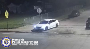 A security‑camera still image shows a white sedan stopped or moving slowly near the corner of a residential street at night. The vehicle’s headlights are on, illuminating the pavement in front. The car is positioned partly on the street and partly near a driveway or curb cut. A grassy area with a posted street sign and a wire fence runs along the left side of the frame. Another vehicle is parked farther up the street on the right. In the lower‑left corner, a Memphis Police Department badge logo appears with the text “Memphis Police Department CRIMESTOPPERS 901‑528‑CASH.”