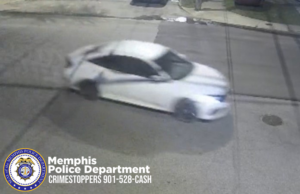 A security‑camera still image shows a white sedan driving through a residential or commercial street at night. The vehicle appears blurred due to motion. The car has dark wheels and tinted windows, and is traveling on a paved road with visible cracks. A curb, some grass, and part of a sidewalk are visible in the background. In the lower-left corner, the Memphis Police Department badge logo and the text “Memphis Police Department CRIMESTOPPERS 901‑528‑CASH” appear.