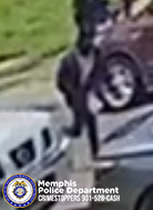 A low‑resolution surveillance‑style image shows a single individual standing on a paved street near the front of a light‑colored vehicle. A second vehicle, which appears to be a darker color, is parked behind the first one and partially visible. The individual is dressed in dark clothing and is positioned near the gap between the two vehicles. In the lower-left corner, a Memphis Police Department logo and the text “Memphis Police Department CRIMESTOPPERS 901‑528‑CASH” are visible.