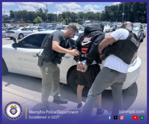 Two individuals wearing tactical vests labeled “Memphis Police Fugitive Unit” are detaining a handcuffed person beside a white Memphis Police Department vehicle in a parking lot filled with numerous parked cars. The detained person is wearing a dark hooded sweatshirt with a large Nike logo on the back and patterned shorts. The scene takes place outdoors under a bright sky with trees and buildings visible in the background. At the bottom of the image, text reads “Memphis Police Department — Established in 1827,” accompanied by the department seal and several social media icons, along with the website joinmpd.com.