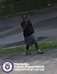 A surveillance‑style, low‑resolution image shows a single person standing on a patch of grass near a street. The person is wearing a dark top, light knee‑length shorts, and light shoes with dark socks. The person is holding one arm up toward the head, though the action is unclear due to image quality. In the lower-left corner, there is a Memphis Police Department logo and text.
