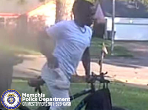 An outdoor residential scene showing a person wearing a light-colored short‑sleeve shirt and light pants standing next to or leaning over a bicycle. One hand rests on the bicycle’s handlebars, and the other hand is near the person’s hip. The background includes grass, a tree trunk, a paved driveway, and several houses with light-colored exteriors. Sunlight creates bright areas on the ground and in the background. In the bottom-left corner of the image, there is a circular badge-like graphic and white text that reads ‘Memphis Police Department CRIMESTOPPERS 901‑528‑CASH.’
