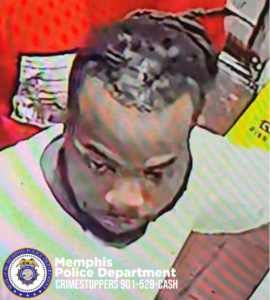  A surveillance-style image shows an individual standing indoors near a counter with red walls in the background. The person is wearing a light-colored top. A stack of folded cloth or towels is visible behind the individual on the left side. On the right side, part of a bright yellow object with black printed text is visible. In the lower-left corner, a circular Memphis Police Department emblem appears, along with white text that reads “Memphis Police Department CRIMESTOPPERS 901-528-CASH.”