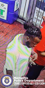 A surveillance camera image shows a person indoors, standing beside another person who is partially visible in a red shirt. The person in the foreground is wearing a plain white T-shirt and has dark hair styled into two braided buns or twists gathered at the back of the head. They have a short beard and are looking downward toward their hands. The floor is covered with reddish-brown tile, and a blue trash or recycling bin is visible in the upper left. Signs are posted on the wall behind them. A watermark at the bottom reads "Memphis Police Department CrimeStoppers 901-528-CASH" alongside a police department badge.