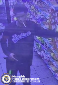 A surveillance-style image taken inside a convenience store shows a person standing in an aisle lined with brightly colored snacks and drinks. The person is wearing a dark long-sleeve shirt with a large “Brookhaven” logo across the chest. A long object resembling a cigarette is in the person’s mouth. One arm is extended outward, and the other hand is positioned near the waist, with fingers pointing downward. The floor consists of square tiles. In the bottom corner, a Memphis Police Department CrimeStoppers badge and the text “Memphis Police Department CRIMESTOPPERS 901-528-CASH” are visible.