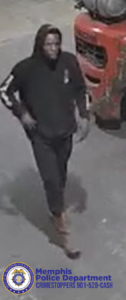 A person is walking on a paved surface near a large red industrial vehicle or forklift. The person is wearing a dark hooded top with light-colored stripes along the sleeves, dark pants, and light-colored footwear. One arm is bent with a hand near the waist, and the other arm hangs down. At the bottom-left corner, there is the Memphis Police Department seal along with text that reads: “Memphis Police Department CRIMESTOPPERS 901-528-CASH.”