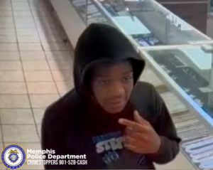 A person wearing a dark hooded sweatshirt stands inside a store with glass display cases containing various small items. The person is positioned near a tiled walkway and is holding one hand raised near the chest area. The setting appears to be a retail environment with bright overhead lighting and multiple display sections. The Memphis Police Department seal and the text “Memphis Police Department CRIMESTOPPERS 901‑528‑CASH” are visible in the lower-left corner of the image.