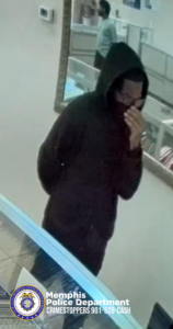 A person wearing a dark hooded sweatshirt stands inside a store with glass display cases containing various items. The person’s hands are raised near the front of the hooded sweatshirt. A large wall mirror reflects another individual standing farther inside the store near additional display counters. The flooring appears to be light-colored tile. In the bottom-left corner of the image, the Memphis Police Department seal is visible along with the text “Memphis Police Department CRIMESTOPPERS 901‑528‑CASH.