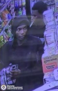 A surveillance-style image taken inside a small convenience store. Two individuals are standing in an aisle lined with brightly colored packaged goods. The individual in the foreground is wearing a dark hoodie with the hood up and is holding an item wrapped in packaging. The individual in the background is wearing a dark shirt and has an object in their mouth that resembles a cigarette. On the right side of the image, shelves display multiple cups labeled “Maruchan Instant Lunch.” At the bottom left corner, a Memphis Police Department badge icon and text read “Memphis Police Department CRIMESTOPPERS 901-528-CASH.