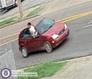 A surveillance-style image shows a small red hatchback car stopped on a residential street near a yellow centerline. A person is leaning out of the vehicle’s front window, positioned on the left side of the car. The background includes a sidewalk, grass, and part of a fence in the lower-left corner. In the bottom-left corner of the image, the Memphis Police Department seal is visible along with text that reads “Memphis Police Department CRIMESTOPPERS 901‑528‑CASH.