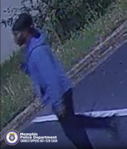 A person wearing a light-colored hooded sweatshirt and dark pants is captured mid‑stride on a paved surface near a grassy area with bushes. The image appears to be from a security camera. In the lower-left corner, there is a Memphis Police Department badge logo with text reading “Memphis Police Department CRIMESTOPPERS 901‑528‑CASH.”