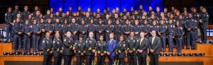 A large group of uniformed law enforcement personnel stands arranged in multiple rows on tiered steps inside an auditorium. Individuals in dress uniforms and formal suits are positioned in the front row, while the remaining personnel wear standard dark uniforms. Police hats are lined up neatly along the edge of the front platform. The background features tall blue-lit panels and stage lighting, creating a formal ceremony or graduation setting.
