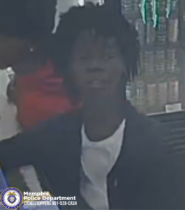 Surveillance-style image taken inside a store. A person with short, textured hair is in the foreground wearing a light-colored shirt under a dark jacket. Another individual in a bright red top is partially visible nearby. Behind them is a refrigerated display case containing cans and bottles, including several cans of an energy drink with a distinctive green claw-mark logo. In the lower-left corner, a Memphis Police Department CrimeStoppers graphic is visible with the text: 'Memphis Police Department CRIMESTOPPERS 901-528-CASH.