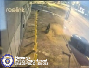A nighttime surveillance camera view shows the side of a commercial building with several yellow bollards lined up along the wall. A dark-colored object or figure stands near the middle of the frame on the pavement. A vehicle is partially visible on the right side of the image, close to the bollards. Streetlights illuminate the surrounding roadway in the background. The bottom left corner contains a Memphis Police Department badge and text: “Memphis Police Department CRIMESTOPPERS 901-528-CASH.