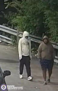 A security-camera image shows two individuals walking outdoors along a paved area next to a metal guardrail. Dense trees and bushes fill the background. The individual on the left is wearing a light-colored hooded sweatshirt with the hood up, dark pants, and light shoes. The individual on the right is wearing a darker hooded sweatshirt with the hood up, patterned shorts, and dark footwear. The front corner of a vehicle is partially visible in the lower left corner. In the bottom-left corner of the image, there is the Memphis Police Department CrimeStoppers logo along with text that reads: "Memphis Police Department CRIMESTOPPERS 901-528-CASH.