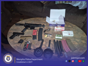 A tabletop is covered with multiple firearms, magazines, and ammunition. Visible items include a rifle-style firearm, several handguns, two drum magazines, multiple extended magazines, standard rifle magazines in different colors, boxed ammunition of various brands, and loose rounds. An open ammunition box sits near the center-right of the table. The table is in a living room with a brown couch in the background. At the bottom of the image, a Memphis Police Department banner reads: “Memphis Police Department — Established in 1827” alongside the department seal and the website “joinmpd.com,” with icons for social media platforms.