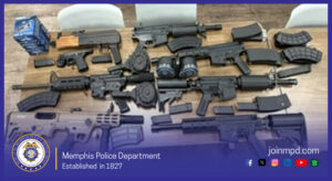 A display of numerous firearms and firearm accessories laid out on a wooden table. The items include several long guns, multiple detachable magazines of various sizes, drum-style magazines, and other weapon components. A box of ammunition is visible in the upper left corner. The table is surrounded by white chairs. The bottom portion of the image contains a Memphis Police Department banner with the department seal on the left, the text “Memphis Police Department – Established in 1827,” and the website “joinmpd.com” along with social media icons on the right. The banner has a blue gradient background.