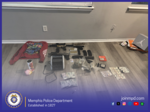 A photograph shows various items laid out on a hardwood floor inside a room with light gray walls and two windows. The items are arranged in rows. On the left side, there is a red hooded sweatshirt with a graphic design on the front. Nearby sits a cup with a colorful pattern and a small fuzzy keychain attached. In the center, there are multiple firearms, magazines, and ammunition lined up, including rifle and handgun components. A large electronic device, possibly a printer or scanner, is positioned near the firearms. To the right, several clear plastic bags containing green leafy material and other substances are spread out. Additional items include small containers, a scale, and stacks of U.S. currency in various denominations arranged neatly. In the far right corner, several shoeboxes and packages are visible. The bottom portion of the image contains a Memphis Police Department banner with its badge, the text “Memphis Police Department – Established in 1827,” and icons for social media alongside the website “joinmpd.com.”