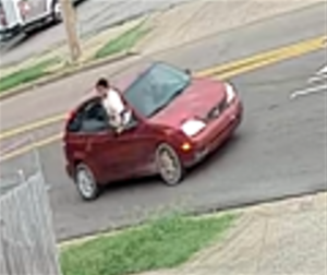 A small red two‑door hatchback car is stopped on a residential street near an intersection. The front wheels are angled as if the car is turning. A person is positioned halfway out of the driver‑side window, leaning out with the upper body extended outside the vehicle. The car appears slightly worn, with visible dirt on the exterior. The background shows a sidewalk, grass, part of a wooden fence, and another vehicle parked farther up the street.