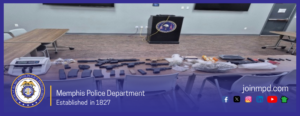 A wide photo shows a conference‑room‑style setting inside a Memphis Police Department facility. In the background, there is a podium with the department’s seal on the front, positioned against a light gray wall with power outlets visible. Large monitors are mounted on the wall above the podium. Two long tables sit on either side of the room. In the foreground, a third table is covered with arranged items. These items include several firearms of different types, numerous magazines, assorted ammunition, various plastic bags containing unidentified materials, multiple small containers, and electronic devices. Chairs are lined up in front of the table. At the bottom of the image, a blue banner displays the Memphis Police Department seal, the text “Memphis Police Department – Established in 1827,” and the website “joinmpd.com.” Beside the website are several small social media icons.