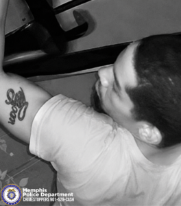A close-up black-and-white surveillance-style image shows the upper body of a person wearing a light-colored short-sleeve shirt while lifting or holding a long object overhead. The person has a tattoo with stylized script lettering on the upper arm. In the lower-left corner, there is a Memphis Police Department CrimeStoppers badge and the text “Memphis Police Department CRIMESTOPPERS 901-528-CASH.” The background includes walls and scattered debris on the ground.