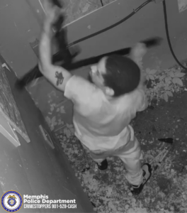 A black‑and‑white surveillance image shows a person inside a narrow, debris‑covered space. The person is holding a long object raised overhead with both arms, appearing to swing it downward. The person is wearing a light‑colored short‑sleeve shirt, light pants, and dark shoes. A visible tattoo is on the person’s upper arm. The walls around the person are damaged, with broken pieces scattered across the floor. Electrical cables or wires run along the upper right side of the wall. In the lower left corner is a Memphis Police Department CrimeStoppers logo with the phone number 901‑528‑CASH.