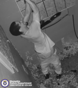 A black‑and‑white surveillance image shows a person standing in a narrow, debris-covered area indoors. The person is lifting a long object overhead with both arms. The person is wearing a light-colored short‑sleeve shirt, light pants, and dark footwear. A tattoo with stylized script lettering is visible on the person’s upper arm. The surrounding walls appear damaged, and broken material is scattered on the ground. In the lower-left corner, there is a Memphis Police Department CrimeStoppers badge and the text “Memphis Police Department CRIMESTOPPERS 901‑528‑CASH.”