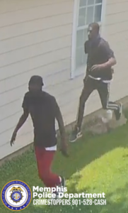 A security‑camera-style image shows two individuals walking outside along the side of a light‑colored house with horizontal siding. One individual is wearing a dark shirt and red pants and is walking closest to the camera. The second individual is wearing a dark shirt and dark pants with a white stripe and is stepping over a low rock or landscaping border near a window. The ground is covered with grass. In the lower-left corner, there is the Memphis Police Department seal and text that reads: “Memphis Police Department CRIMESTOPPERS 901‑528‑CASH.”