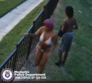 The image shows two people outdoors beside a black metal fence. In the foreground, a person wearing a light-colored bikini and a pink head covering is leaning against the fence. Behind them, a shirtless person in gray shorts is walking away across a grassy area. A sidewalk runs along the left side of the image. A “Memphis Police Department” watermark appears in the lower-left corner.