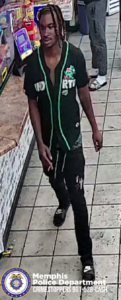 A surveillance-style image inside a convenience store shows one individual walking on a tiled floor near a counter. The individual is wearing a dark short-sleeved top with bright green trim and printed graphics on the front, along with dark pants that have printed designs on the upper thigh area. Footwear appears dark with light markings. The individual's hair has multiple light-colored sections. Behind the individual, part of another person is visible wearing long pants and open-toe footwear. To the left, shelves and displays hold various brightly colored packaged products. At the bottom of the image, a logo for the Memphis Police Department appears, along with text that reads: “Memphis Police Department CRIMESTOPPERS 901-528-CASH.”