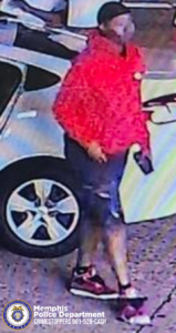 A surveillance-style image showing an individual standing beside a white sedan in a parking area. The individual is wearing a bright red hooded jacket, dark shorts with a torn or distressed area, dark footwear with red accents, and a dark face covering or mask. The individual is holding an object in one hand. The ground surface appears textured like concrete or pavement. The Memphis Police Department emblem and the text “Memphis Police Department CRIMESTOPPERS 901‑528‑CASH” are visible in the lower-left corner of the image.