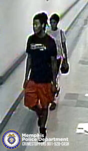 A surveillance-style image shows two individuals walking down a long, indoor hallway. The person in front is wearing a dark short-sleeved shirt and bright knee-length shorts, carrying something in one hand. The person behind is wearing a light-colored top and carrying a shoulder bag. The hallway has a tiled floor and light-colored walls. In the bottom left corner of the image, there is a Memphis Police Department badge and text that reads: “Memphis Police Department CRIMESTOPPERS 901-528-CASH.”