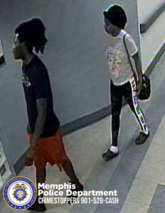 A surveillance-style image shows two individuals walking down a hallway with light-colored flooring and walls. The individual in front is wearing a dark short-sleeved shirt and bright knee-length shorts, walking toward the left side of the image. The individual behind is wearing a light-colored top with a graphic design, dark pants with a vertical stripe down the side, and is carrying a bag over one shoulder. The flooring has a geometric pattern and the hallway contains handrails along the walls. In the lower-left corner, a Memphis Police Department badge is visible next to text that reads: “Memphis Police Department CRIMESTOPPERS 901-528-CASH.”
