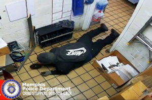 A surveillance-style image shows a person lying face‑down on a tiled floor inside a small room with white brick walls. The person is wearing dark clothing, including a hoodie with a large Nike swoosh logo across the back and a face covering. One arm is extended forward, and the person appears to be holding an object. Around the room are various shelves, boxes, and cleaning supplies. In the lower-left corner, there is a Memphis Police Department seal and text that reads “Memphis Police Department CRIMESTOPPERS 901‑528‑CASH.”